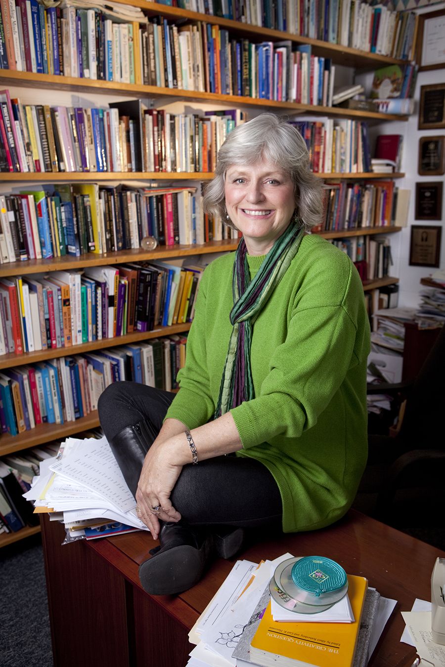 Bonnie Cramond sits cross-legged on top of a desk cluttered with stacks of paper, books and compact discs. Behind her, 7 bookshelves stuffed full of books of every size and color fill the frame. She is wearing a kelly green tunic and striped scarf in tones of green and purple.